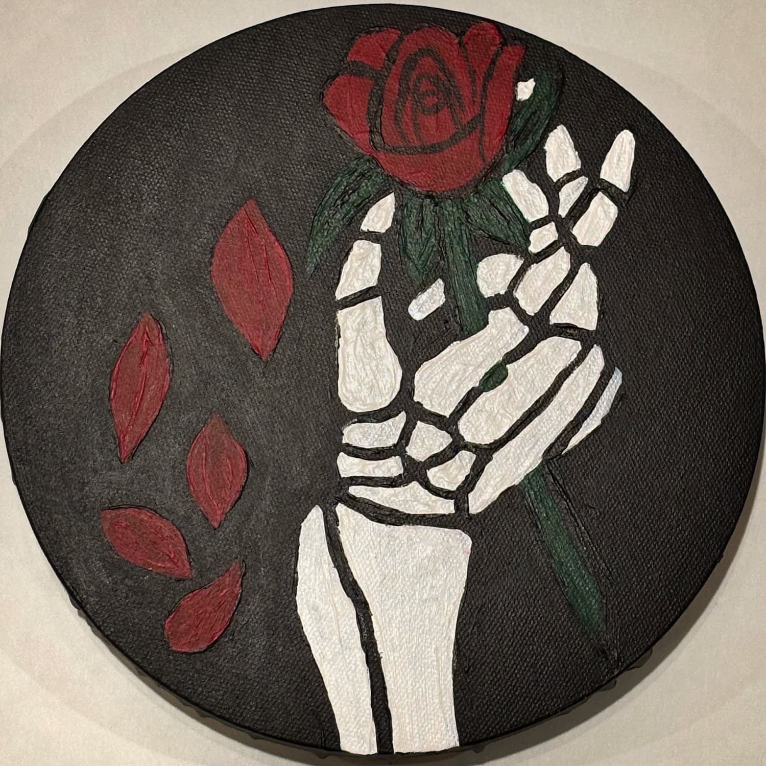 Round acrylic painting of a skeleton hand holding a red rose, off-white background